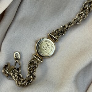May include: A gold-toned watch bracelet with a round watch face. The watch face has the letters "CC" inside a circle. The bracelet is made of a thick, rope-like chain. The watch is on a beige surface.