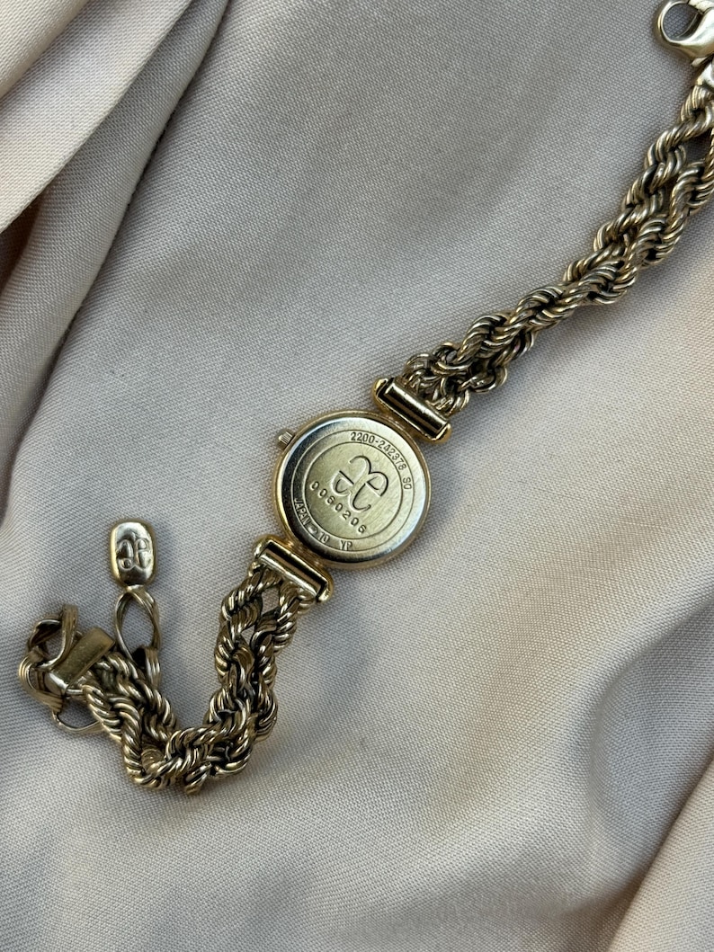 May include: A gold-toned watch bracelet with a round watch face. The watch face has the letters "CC" inside a circle. The bracelet is made of a thick, rope-like chain. The watch is on a beige surface.