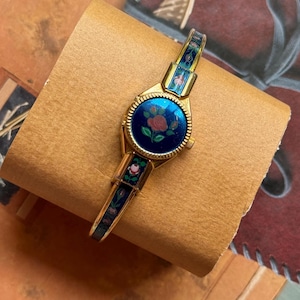 Puede incluir: Un reloj de pulsera rígida en tono dorado con una esfera redonda de esmalte azul profundo y un motivo central de rosa roja. La banda está decorada con paneles de esmalte azul a juego y pequeños patrones florales, con un bisel dorado estriado.