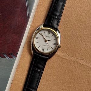 Puede incluir: Un reloj de pulsera Seiko Lucent con esfera blanca, números romanos negros, bisel dorado y caja plateada. El reloj cuenta con una correa de cuero negro con un patrón texturizado en relieve y una corona de color dorado en el lateral.