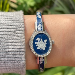 Op de afbeelding: Een vintage zilverkleurig horloge met een blauwe wijzerplaat en een wit bloemmotief. Het horloge heeft een rechthoekige wijzerplaat met een wit bloemmotief en is omgeven door kleine, heldere kristallen. De horlogeband is blauw met witte bloemaccenten.