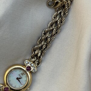 May include: A gold-toned wristwatch with a mother-of-pearl face and a braided chain band. The watch features a round face with the brand name and is adorned with red and clear gemstones.