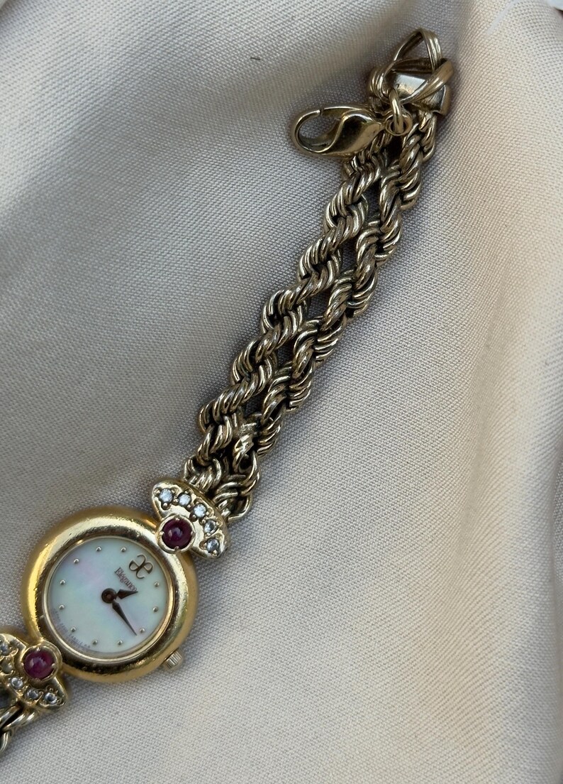May include: A gold-toned wristwatch with a mother-of-pearl face and a braided chain band. The watch features a round face with the brand name and is adorned with red and clear gemstones.