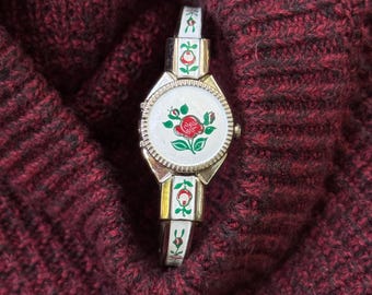 May include: A silver-tone wristwatch featuring a white dial with a central red rose and green leaf illustration. The metal band is decorated with white enamel panels, each displaying a small red rosebud and green leaves, matching the watch face design.