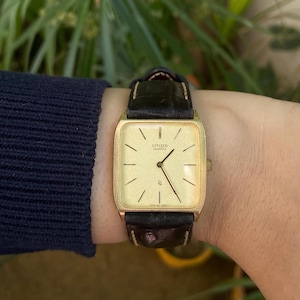 Könnte beinhalten: Eine goldfarbene Citizen Quartz-Uhr mit rechteckigem Zifferblatt und schwarzem Lederarmband. Das Zifferblatt der Uhr ist hellgoldfarben mit schwarzen Stundenmarkierungen und Zeigern. Die Uhr befindet sich an einem Handgelenk.