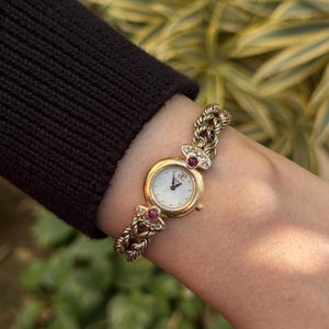Puede incluir: Un reloj de pulsera dorado con esfera blanca y agujas doradas. El reloj tiene una correa trenzada y está adornado con pequeñas piedras preciosas rojas. El reloj se lleva en la muñeca, con una manga negra en el fondo.