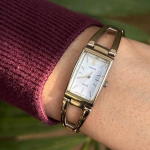 May include: A gold Citizen Clelia watch with a rectangular face and a white mother-of-pearl dial. The watch has a gold band with an open design. The watch is displayed on a wrist, with a burgundy sleeve in the background.