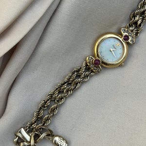 May include: A gold-toned wristwatch with a round face and a mother-of-pearl dial. The watch features a gold bezel, red gemstones, and a braided chain-link band. The watch is a vintage style.