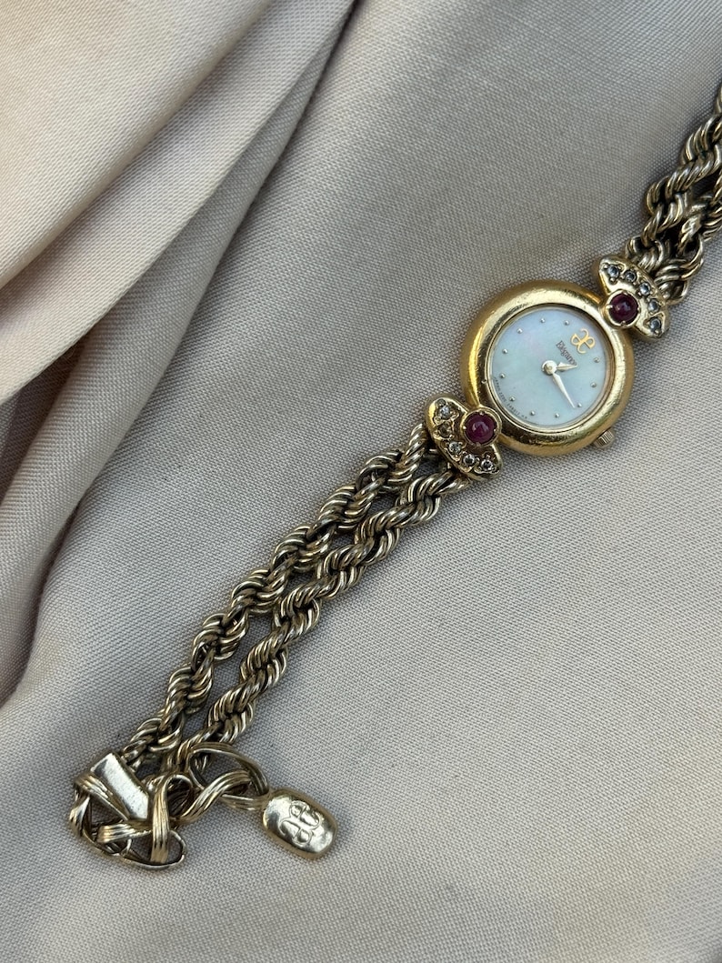 May include: A gold-toned wristwatch with a round face and a mother-of-pearl dial. The watch features a gold bezel, red gemstones, and a braided chain-link band. The watch is a vintage style.