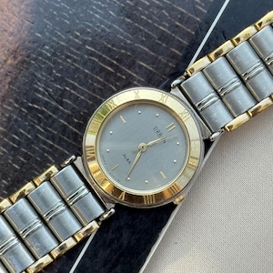 May include: A vintage Alba Urbn watch with a silver face and gold-tone bezel and hands. The watch features Roman numeral hour markers and a two-tone silver and gold-tone metal strap. The watch is displayed on a patterned surface.