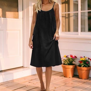 May include: A black midi dress with spaghetti straps. The dress has a loose, flowing silhouette and falls to mid-calf. The model is standing on a wooden porch with potted flowers in the background.