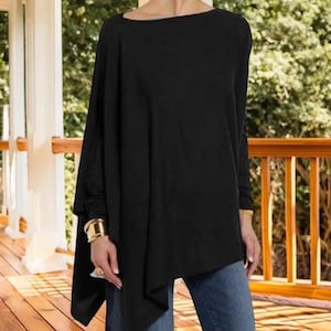 May include: A black long-sleeve top with a boat neck and an asymmetrical hemline. The top has a loose, flowing design. The sleeves are long and fitted at the wrists. The model is wearing blue jeans. The background is a wooden deck with a natural setting.