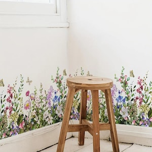 May include: A wooden stool stands in front of a white wall adorned with a floral decal. The decal features a colorful array of wildflowers and butterflies in shades of pink, purple, blue, and yellow. The stool has a round seat and three legs.