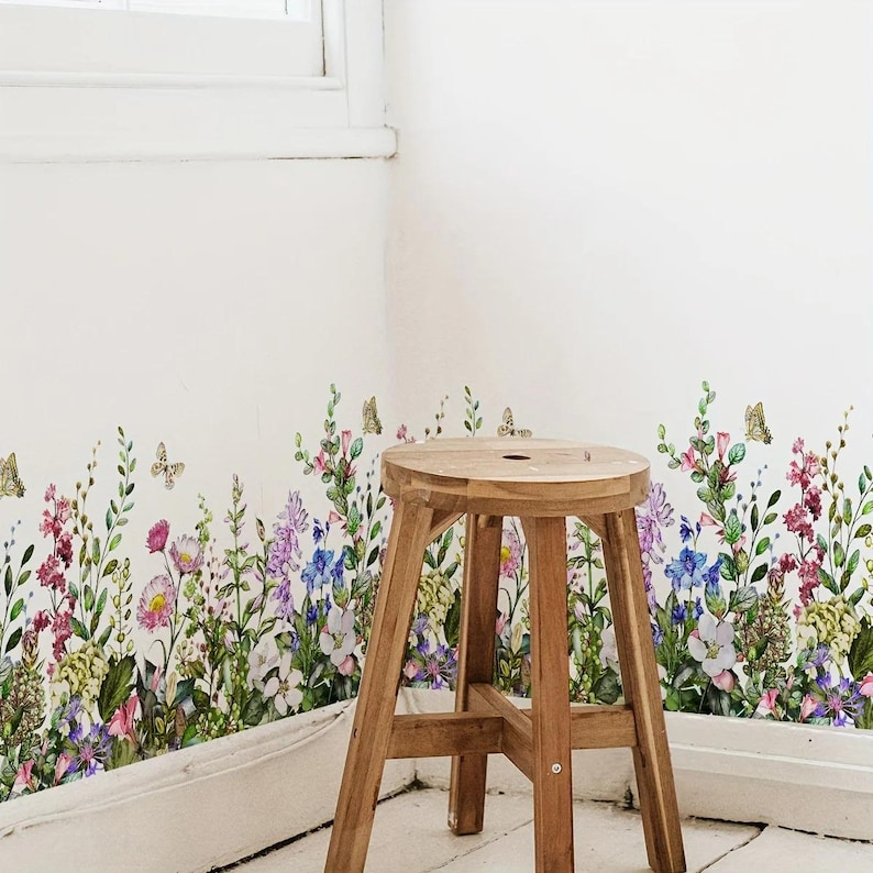 May include: A wooden stool stands in front of a white wall adorned with a floral decal. The decal features a colorful array of wildflowers and butterflies in shades of pink, purple, blue, and yellow. The stool has a round seat and three legs.