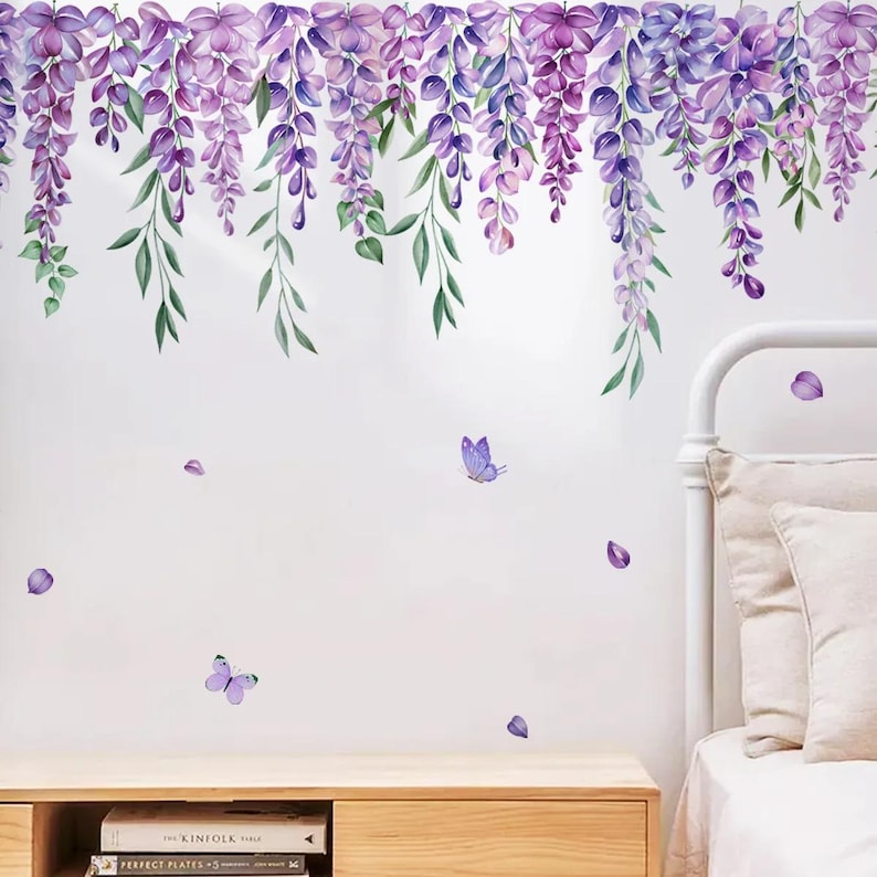 May include: A wall decal featuring watercolor wisteria flowers in shades of purple and green. The design includes hanging flower clusters, green leaves, and scattered purple butterflies and petals. The decal is applied to a white wall.