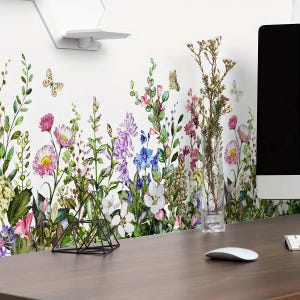 May include: A white wall decorated with a colorful floral decal featuring various flowers and butterflies. The decal includes pink, purple, blue, and white flowers with green foliage. A black geometric object sits on a wooden desk.