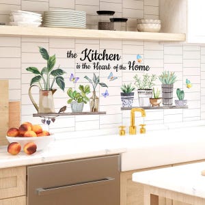 May include: A kitchen wall decal with the text "the Kitchen is the Heart of the Home". The decal features watercolor illustrations of potted plants, butterflies, and a bird. The kitchen has a white sink, a gold faucet, and wooden shelves.