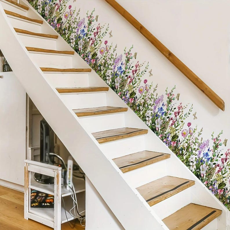 May include: White staircase with wooden steps and a wooden handrail. A floral wall decal with colorful flowers and butterflies decorates the wall. The stairs lead upwards, with a small shelf and cabinet visible below.
