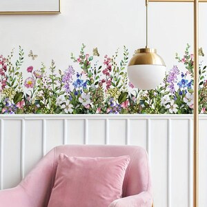 May include: A floral wall decal featuring a vibrant array of wildflowers in shades of pink, purple, blue, and white, with green foliage and butterflies. The decal is applied above a white panel wall and a pink armchair.
