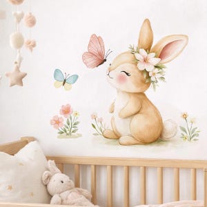 May include: A wall decal featuring a light brown bunny with pink cheeks and a floral crown, accompanied by a pink butterfly, a blue and yellow butterfly, and small pink and yellow flowers, positioned above a wooden crib with a plush toy.