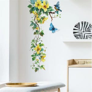 Op de afbeelding: Muursticker met een aquarelontwerp van gele bloemen, groene bladeren, een blauwe vogel en een blauwe vlinder. De bloemstukken vallen langs de muur naar beneden en creëren een door de natuur geïnspireerd decoratief element. De sticker is aangebracht op een witte muur.