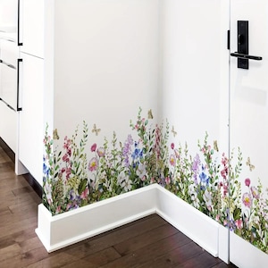May include: A floral wall decal featuring a colorful wildflower meadow design. The decal includes various flowers in shades of pink, purple, blue, and yellow, with green foliage and butterflies. The decal is applied to a white wall, creating a vibrant and natural aesthetic.