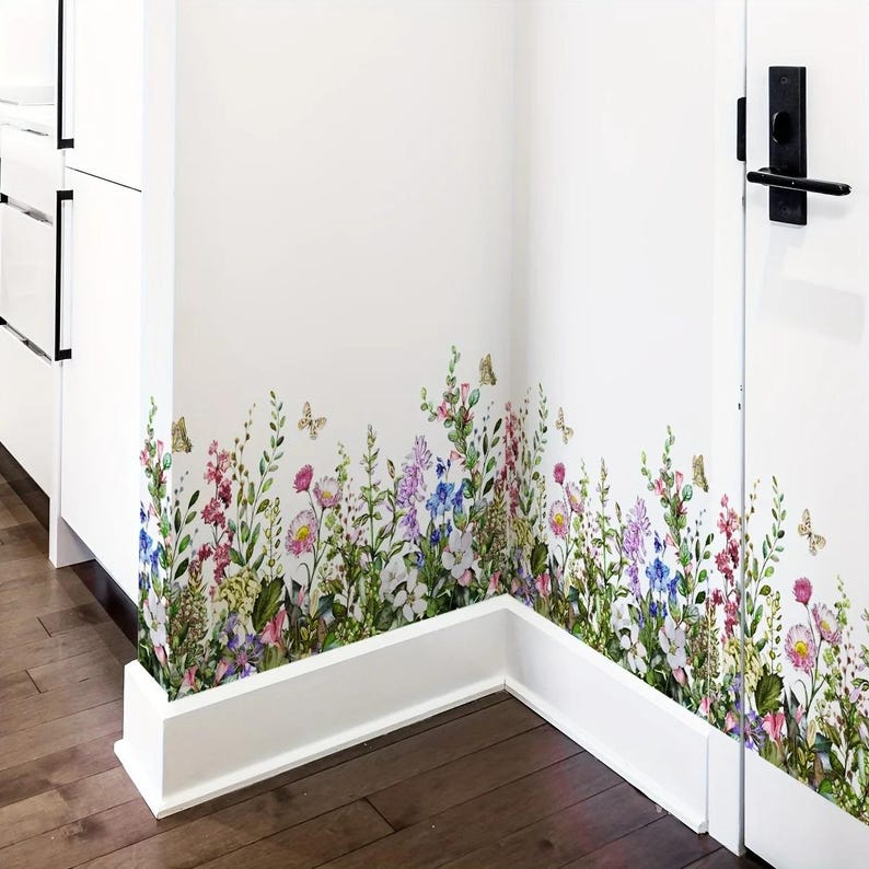 May include: A floral wall decal featuring a colorful wildflower meadow design. The decal includes various flowers in shades of pink, purple, blue, and yellow, with green foliage and butterflies. The decal is applied to a white wall, creating a vibrant and natural aesthetic.