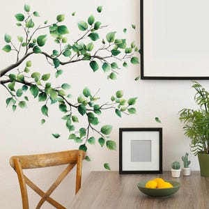 May include: A wall decal featuring a watercolor design of tree branches with green leaves. The branches extend across a white wall, with some leaves appearing to fall. A wooden chair and table are in the foreground, with a framed picture and a bowl of lemons.