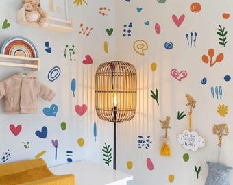 May include: A nursery room with a white wall decorated with colorful painted shapes, including hearts, circles, and flowers. A wooden lamp, a changing table, and a shelf with a teddy bear and rainbow decor are visible.