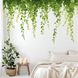 May include: Green leafy wall decals in a bedroom setting, arranged to resemble hanging vines. A potted plant, candles, and a bed with white linens are also visible.