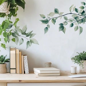 Op de afbeelding: Een thuiskantoor met een houten bureau, boeken en potplanten. Groene, bladerrijke takken zijn op de witte muur geschilderd. Een vetplant in een geweven pot en een hangplant dragen bij aan de natuurlijke esthetiek.