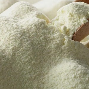 Puede incluir: Primer plano de un montón de polvo amarillo claro, posiblemente un producto alimenticio. Una cuchara de madera está parcialmente insertada en el polvo, sosteniendo una porción. El polvo tiene una textura fina y granulada.