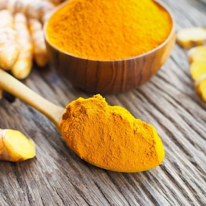 Puede incluir: Una cuchara de madera llena de polvo de cúrcuma de color naranja brillante, junto a un cuenco de madera con polvo y raíces frescas de cúrcuma. La imagen muestra la especia y su forma natural sobre una superficie de madera rústica.