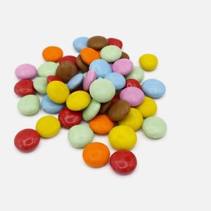 May include: A collection of small, round sweets in a variety of colours: red, orange, yellow, green, blue, pink, and brown. The sweets are scattered on a white background, creating a bright and cheerful image.