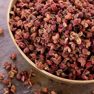 May include: A wooden bowl brimming with dried, reddish-brown Sichuan peppercorns. Some peppercorns are scattered around the bowl on a dark surface. The peppercorns have a textured, open appearance.