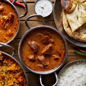 May include: A selection of Indian dishes, including curries, rice, and naan bread, presented in metal bowls. A small dish of yogurt and red chillies are also visible. The food is arranged on a wooden table, creating a colourful display.