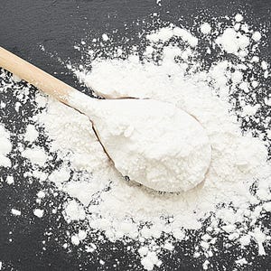 May include: A wooden spoon filled with white flour rests on a dark gray surface, surrounded by scattered flour. The spoon is light brown, and the flour creates a textured, contrasting visual.