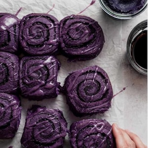 May include: An overhead shot shows purple cinnamon rolls drizzled with white glaze. The rolls are on parchment paper, with two small glass bowls containing purple and dark brown sauces. A hand is visible on the right side of the image.