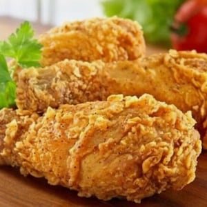 May include: Close-up of golden-brown fried chicken drumsticks, with crispy, textured skin. Garnished with fresh green parsley. The chicken is on a wooden surface, with a tomato and lettuce in the background. A delicious and appetizing meal.