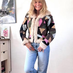 May include: A jacket featuring a geometric patchwork pattern in shades of brown, black, pink, and light blue, accented with cream-colored faux fur trim along the collar, front opening, and cuffs, paired with light blue denim jeans and brown boots.