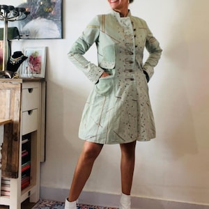 May include: A light green knee-length coat featuring a high collar, a front button closure, and two front pockets. The garment displays a patchwork-style design with visible stitching details and a subtle pattern on the front panel.