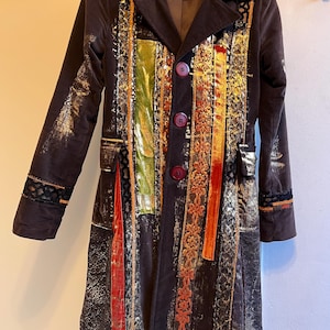 Può includere: Un cappotto in velluto marrone scuro con strisce verticali colorate oro, rosso, verde e arancione. Il cappotto ha un colletto a lancia, tre grandi bottoni e finiture decorative sulle maniche. Il cappotto è appeso a una gruccia di legno.
