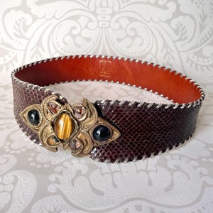 Peut inclure: Large ceinture marron avec un motif peau de serpent et une boucle décorative dorée. La boucle présente une pierre œil de tigre centrale, flanquée de pierres noires et de petites gemmes. Les bords de la ceinture sont cousus avec du fil argenté.