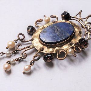 Peut inclure: Broche de style vintage avec une pierre ovale bleu foncé au centre, entourée d'un cadre en métal doré martelé. La broche comporte des perles pendantes et de petites pierres précieuses rouge foncé, créant un design orné et élégant.