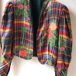 Può includere: Giacca corta vintage con un vivace motivo a quadri in rosso, verde, giallo e blu. La giacca presenta un design floreale di rose rosse e foglie verdi. La giacca è appesa a una gruccia nera.