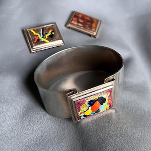 May include: A silver-toned metal cuff bracelet featuring a rectangular centrepiece with a multi-coloured enamel inlay of red, orange, yellow, blue, and black abstract shapes, accompanied by two matching rectangular metal pieces with similar designs.