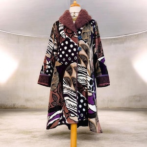 Pode incluir: Um casaco de manga comprida com um padrão de retalhos multicolorido em tons de roxo, castanho, preto e branco. O casaco tem uma gola de pelo sintético malva texturizado, dois botões azuis grandes na frente e punhos com um rebordo roxo escuro.
