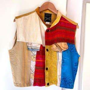Puede incluir: Un chaleco patchwork con una variedad de telas en dorado, rojo, azul y crema. El chaleco tiene cuello, botones negros y una etiqueta que dice "ILLICIT KRAFT". Cuelga de una percha de madera.