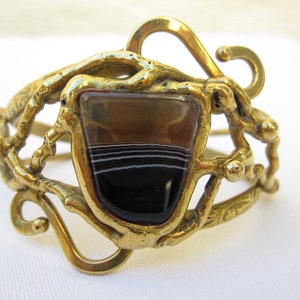 May include: A gold-toned bracelet featuring a large, rectangular, banded agate stone set in the centre. The bracelet has a unique, organic design with intricate details.