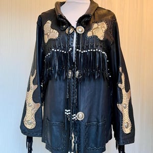 May include: A black leather jacket featuring tan snakeskin-patterned patches on the chest and sleeves. The jacket includes long black leather fringe, braided leather trim, silver-toned metal concho buttons, and white bead accents along the front.